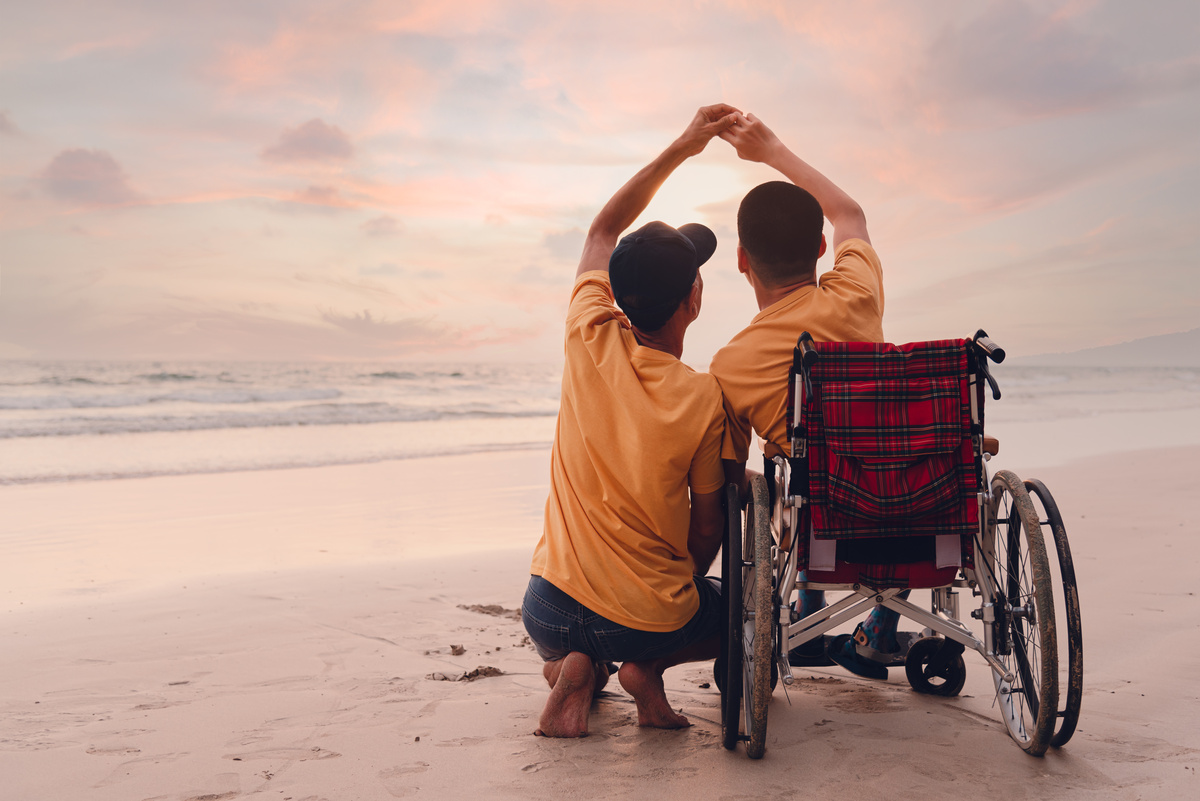 Un père et son fils en fauteuil roulant, de dos, se tiennent la main en observant le coucher de soleil sur la plage