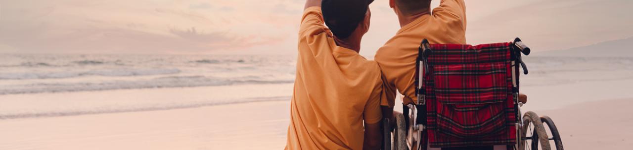 Un père et son fils en fauteuil roulant, de dos, se tiennent la main en observant le coucher de soleil sur la plage